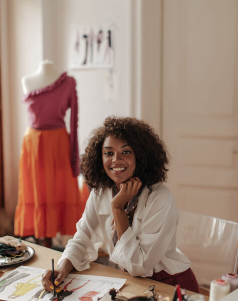 Brown-eyed beautiful curly dark-skinned woman in stylish white blouse sits at desk, designs stylish clothes and smiles.
